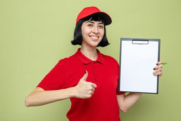 Smiling young caucasian delivery girl holding clipboard and thumbing up 