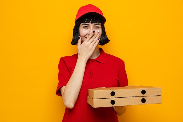 Sorridente giovane ragazza delle consegne caucasica che tiene in mano scatole per pizza e si mette la mano sulla bocca