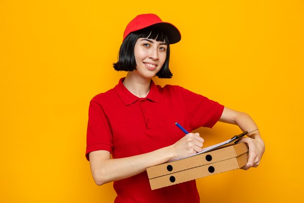 Smiling young caucasian delivery girl holding pizza boxes and writing on clipboard