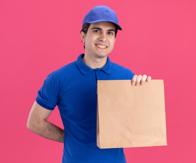 Улыбающийся молодой кавказский курьер в синей форме и кепке держит бумажный пакет, держа руку за спиной