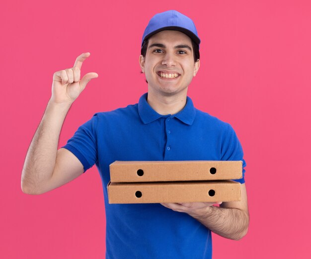 smiling young caucasian delivery man in blue uniform and cap holding pizza packages  showing small amount gesture isolated on pink wall