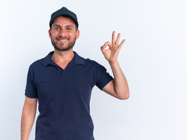 Smiling young caucasian delivery man in blue uniform and cap looking at camera doing ok sign isolated on white wall with copy space