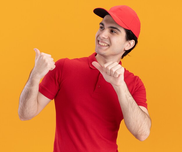 Smiling young caucasian delivery man in blue uniform and cap looking and pointing at side isolated on orange wall