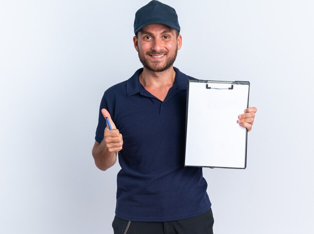 Smiling young caucasian delivery man in blue uniform and cap showing clipboard and thumb up holding pen 