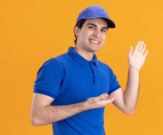 Smiling young caucasian delivery man in blue uniform and cap showing empty hand pointing at it 