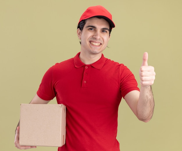 Free photo smiling young caucasian delivery man in red uniform and cap holding cardbox  showing thumb up isolated on olive green wall