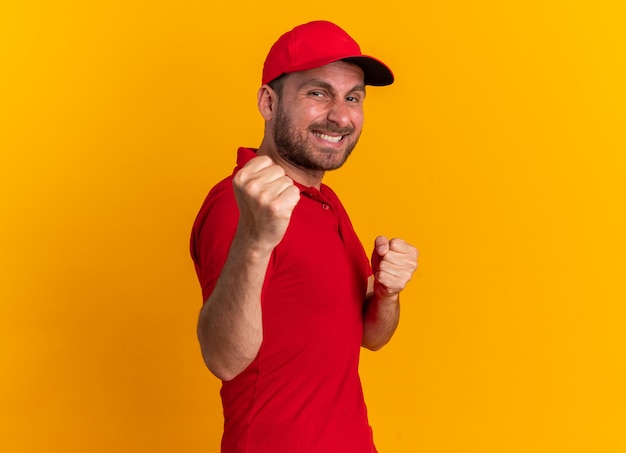 Улыбающийся молодой кавказский доставщик в красной форме и кепке, стоящий в профиле, глядя в камеру, делает боксерский жест, изолированный на оранжевой стене с копией пространства