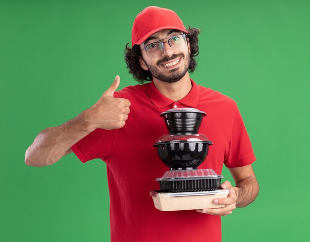 Улыбающийся молодой кавказский курьер в красной форме и кепке в очках держит бумажный пакет с едой и контейнеры для еды, показывая большой палец вверх