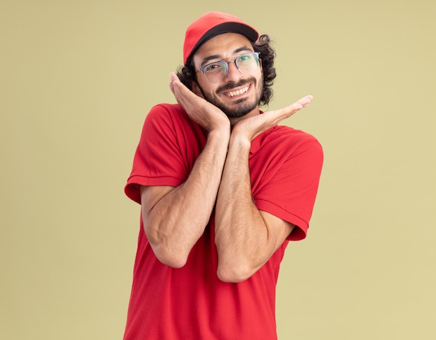 Sorridente giovane fattorino caucasico in uniforme rossa e berretto con gli occhiali tenendo le mani vicino al viso isolato su parete verde oliva con spazio di copia