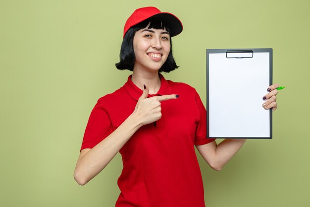 Smiling young caucasian delivery woman holding and pointing at clipboard 