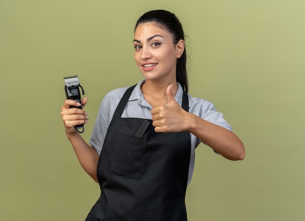 Free photo smiling young caucasian female barber wearing uniform holding hair clippers showing thumb up isolated on olive green wall