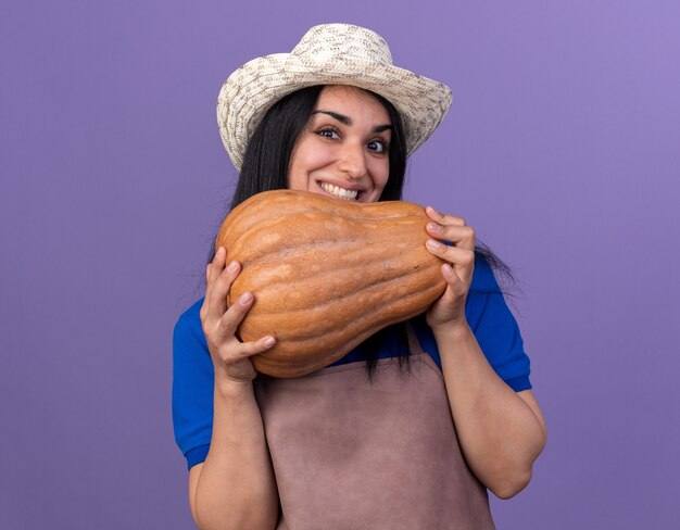 Smiling young caucasian gardener girl wearing uniform and hat holding butternut pumpkin  isolated on purple wall