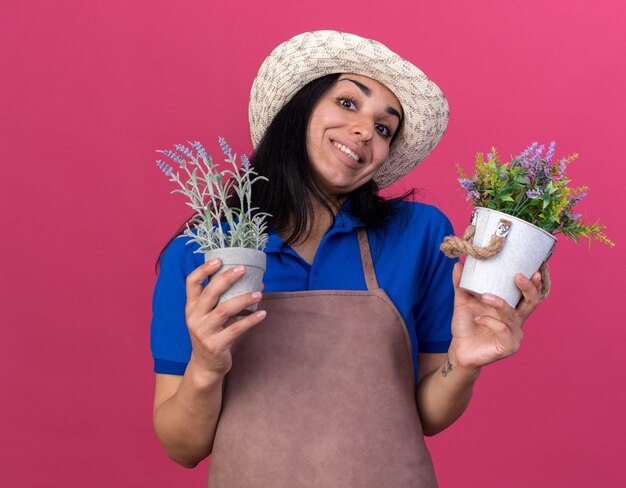 ピンクの壁に分離された植木鉢を保持している制服と帽子を身に着けている若い白人の庭師の女の子の笑顔