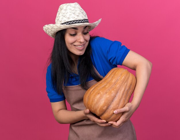 Smiling young caucasian gardener woman wearing uniform and hat holding and looking at butternut pumpkin