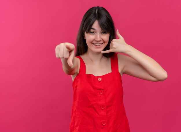 Smiling young caucasian girl does call gesture and points at camera on isolated pink background with copy space