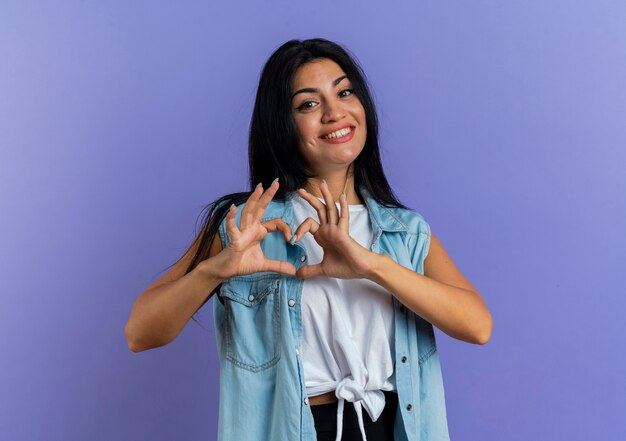 Smiling young caucasian girl gesturing heart hand sign isolated on purple background with copy space