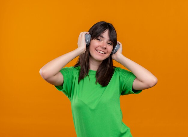 Smiling young caucasian girl in green shirt holds headphones on head on isolated orange background