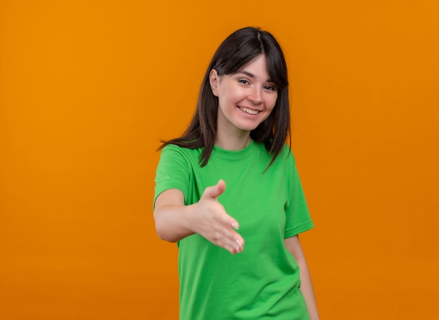 Smiling young caucasian girl in green shirt holds out hand and looks at camera on isolated orange background with copy space