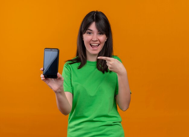 Smiling young caucasian girl in green shirt holds phone and points at phone on isolated orange background