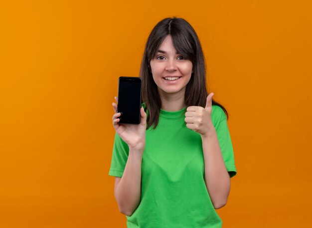 Smiling young caucasian girl in green shirt holds phone and thumbs up on isolated orange background