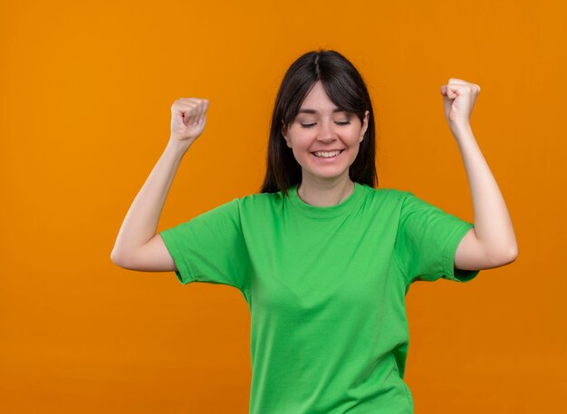 Smiling young caucasian girl in green shirt raises fists up and looks down on isolated orange background with copy space