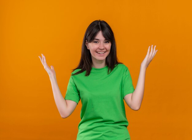 Smiling young caucasian girl in green shirt raises hands and looks at camera on isolated orange background