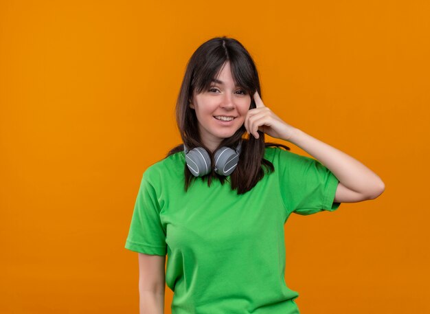 Smiling young caucasian girl in green shirt with headphones puts hand on head on isolated orange background with copy space