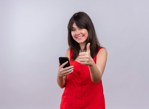 Smiling young caucasian girl holding phone and doing thumbs up gesture looking at camera on isolated white background with copy space