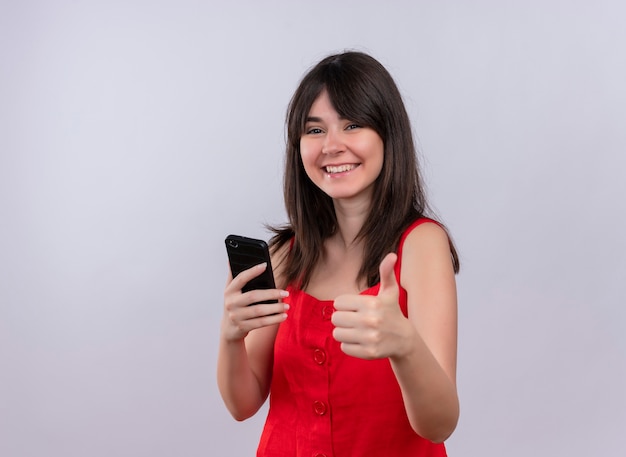 Smiling young caucasian girl holding phone and doing thumbs up gesture looking at camera on isolated white background with copy space