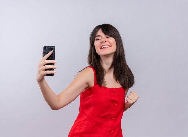 Smiling young caucasian girl holding phone and raising fist looking at camera on isolated white background