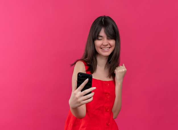 Smiling young caucasian girl holds phone and raises fist up looking at phone on isolated pink background with copy space