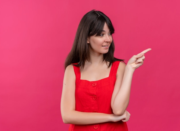 Smiling young caucasian girl points to the side and looks to the side on isolated pink background with copy space