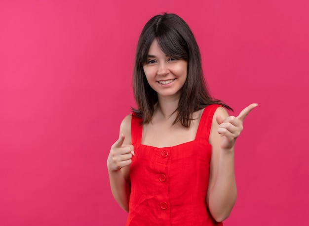 Smiling young caucasian girl points to the side with both hands on isolated pink background with copy space