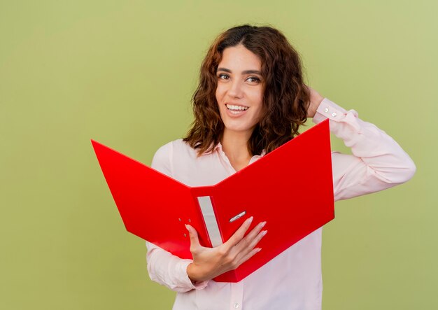 Smiling young caucasian girl puts hand on head behind holding file folder and looking at camera isolated on green background with copy space