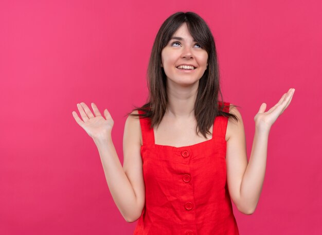 Smiling young caucasian girl raises both hands up and looking up on isolated pink background with copy space