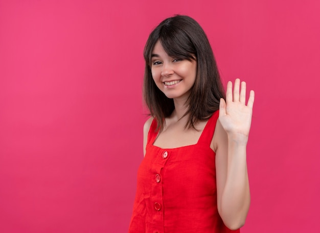 Smiling young caucasian girl raises hand up and looking at camera on isolated pink background with copy space