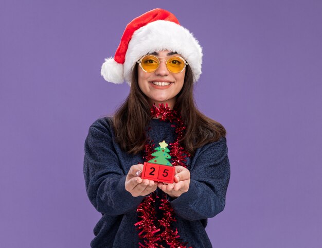 Smiling young caucasian girl in sun glasses with santa hat and garland around neck holding christmas tree ornament isolated on purple wall with copy space