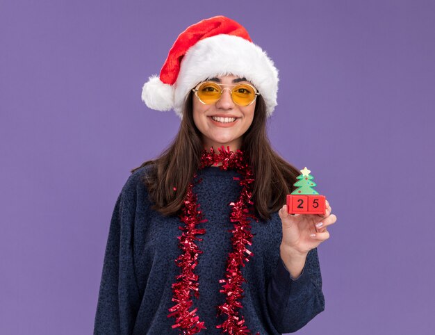 Smiling young caucasian girl in sun glasses with santa hat and garland around neck holds christmas tree ornament isolated on purple wall with copy space