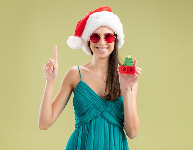 산타 모자 크리스마스 트리 장식을 들고 가리키는 태양 안경에 웃는 젊은 백인 여자