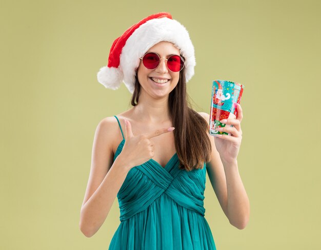 Smiling young caucasian girl in sun glasses with santa hat holding and pointing at paper cup 