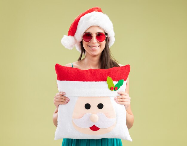 Smiling young caucasian girl in sun glasses with santa hat holding santa pillow 