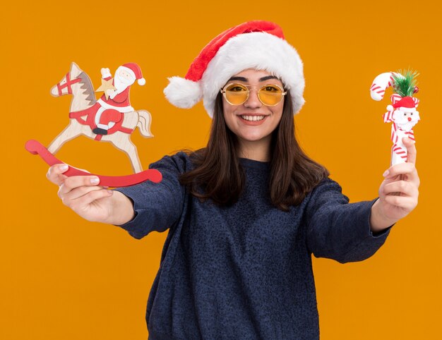 Smiling young caucasian girl in sun glasses with santa hat holding santa on rocking horse decoration and candy cane isolated on orange wall with copy space