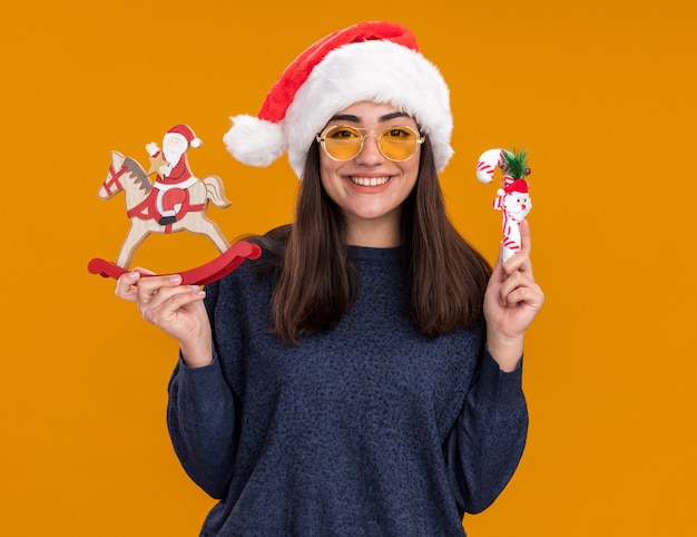 Smiling young caucasian girl in sun glasses with santa hat holds santa on rocking horse decoration and candy cane isolated on orange wall with copy space