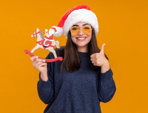 산타 모자와 태양 안경에 웃는 젊은 백인 여자는 흔들 목마 장식에 산타를 보유하고 복사 공간이 오렌지 벽에 고립 엄지 손가락