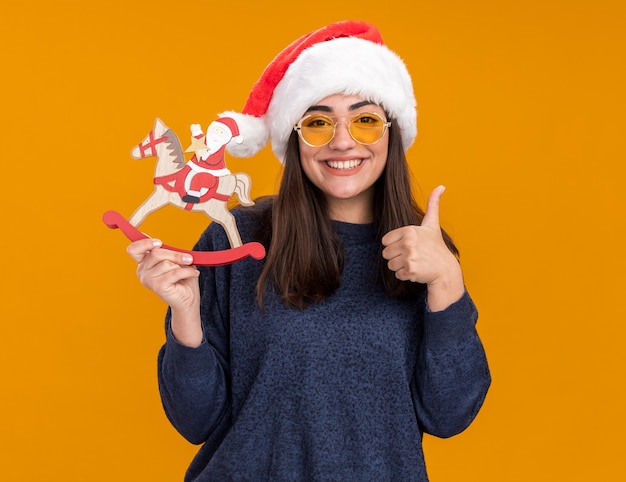Улыбающаяся молодая кавказская девушка в солнцезащитных очках в шляпе санта-клауса держит Санта-Клауса на украшении лошади-качалки и показывает палец вверх, изолированную на оранжевой стене с копией пространства