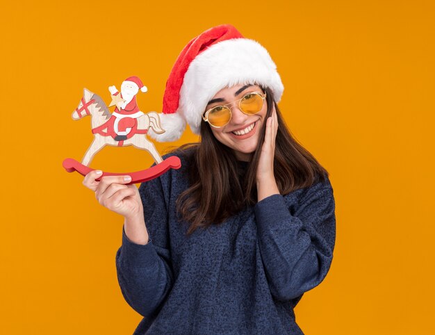 Smiling young caucasian girl in sun glasses with santa hat puts hand on face and holds santa on rocking horse decoration isolated on orange wall with copy space