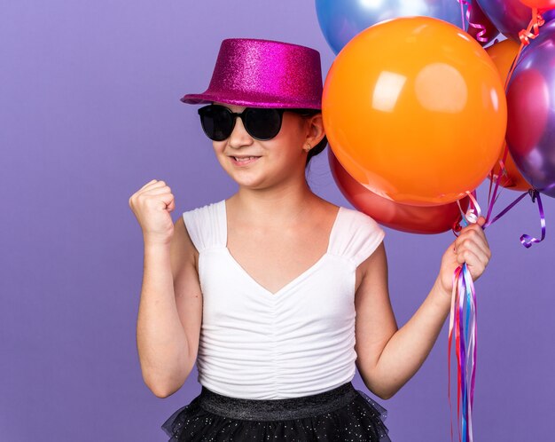 Free photo smiling young caucasian girl in sun glasses with violet party hat holding helium balloons and keeping fist up isolated on purple wall with copy space