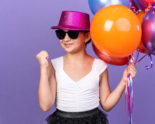 smiling young caucasian girl in sun glasses with violet party hat holding helium balloons and keeping fist up isolated on purple wall with copy space