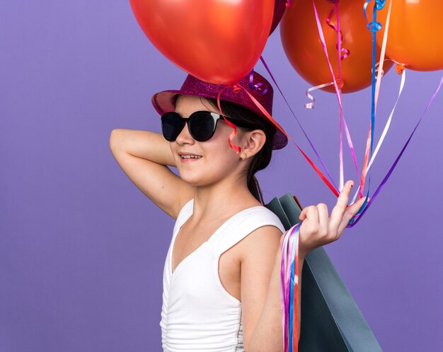 smiling young caucasian girl in sun glasses with violet party hat holding helium balloons and shopping bags on shoulder isolated on purple wall with copy space