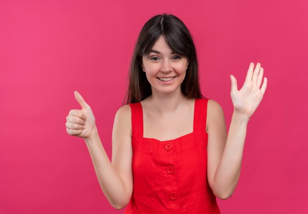 Smiling young caucasian girl thumbs up and raises hand on isolated pink background